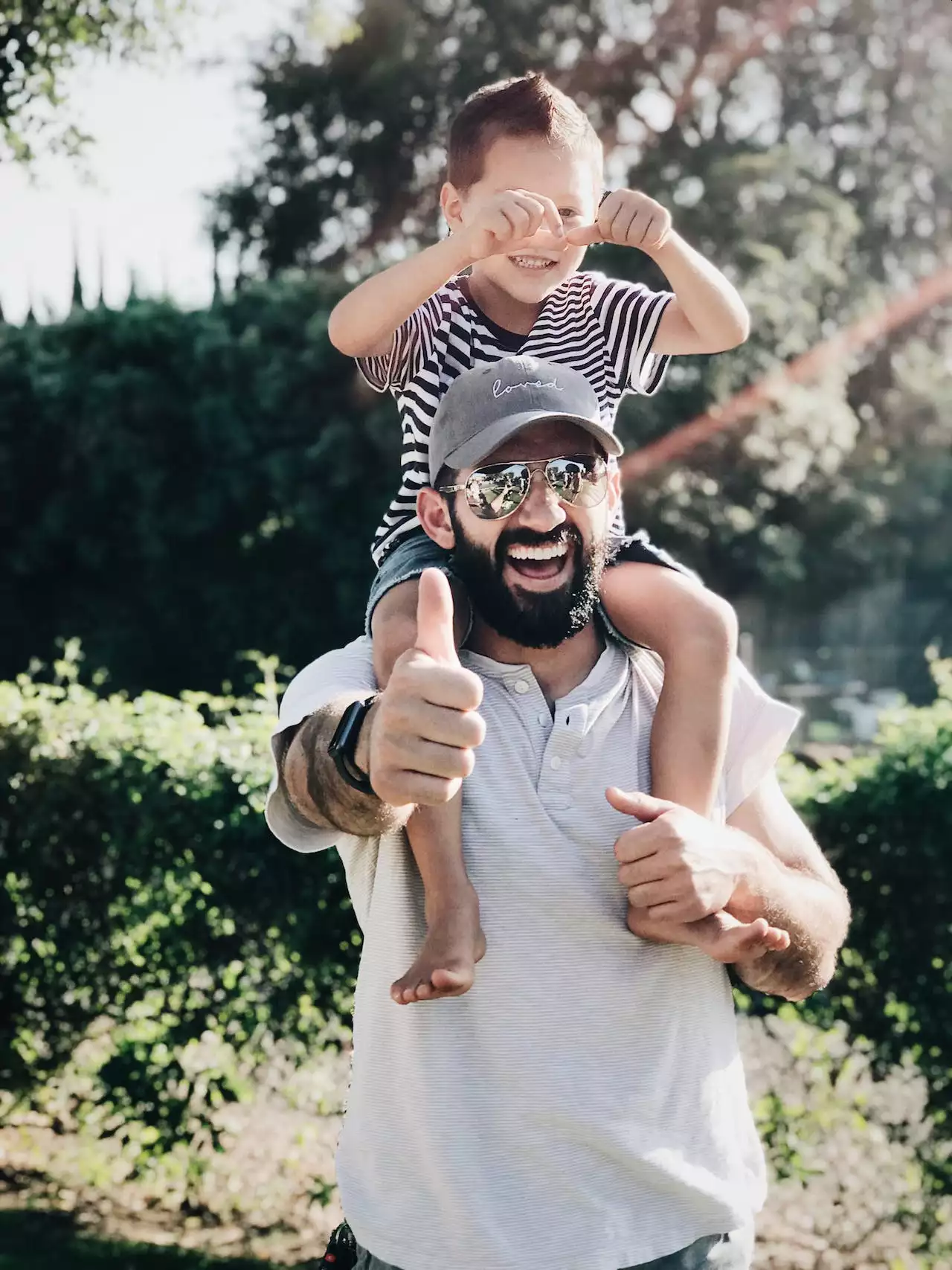 A smiling man outdoors carrying a child on his shoulders